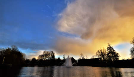 stunning orange and blue sunset with spraying fountain and tree silhouettes reflecting in the pond waterの写真素材