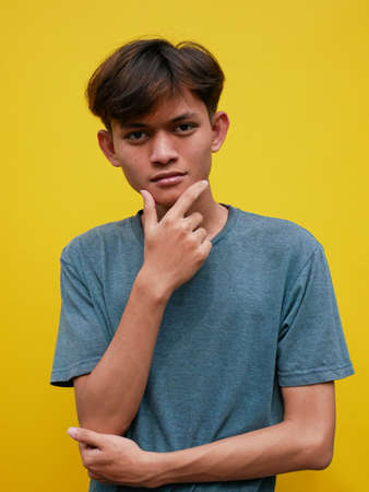 Good offer. Portrait of pensive Asian man looking into empty space, touching his chin, over yellow studio background. teenage boy who thinks and makes decisionsの写真素材