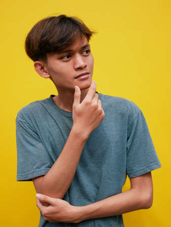 Good offer. Portrait of pensive Asian man looking into empty space, touching his chin, over yellow studio background. teenage boy who thinks and makes decisionsの写真素材