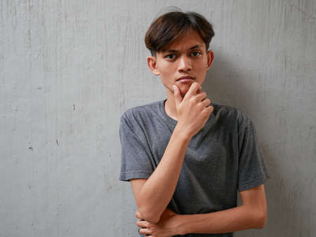 Portrait of pensive Asian man looking into empty space, touching his chin, on gray background. teenage boy who thinks and makes decisionsの写真素材