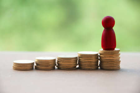 Red wooden doll standing on stack of gold coins. Business concept and copy spaceの写真素材