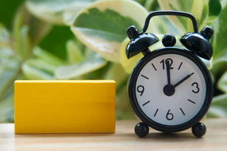 Black alarm clock and blank yellow wooden block isolated on blurred nature background. The clock set at 2 o'clock. Copy space.の写真素材
