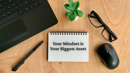 Business motivational text on notebook - Your mindset is your biggest asset. With pen, plant, mouse, laptop and glasses background.の写真素材