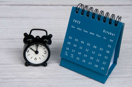October 2022 blue color desk calendar on wooden desk with copy space and analog alarm clock