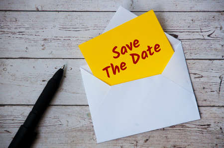 Save the date text on yellow notepad in an envelope with pen and wooden desk background.