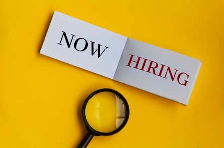 Now hiring text on notepad with magnifying glass on yellow background. Employment conceptの写真素材