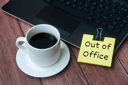 Coffee cup and laptop on wooden table with yellow notepad with text - Out of office. Business conceptの写真素材