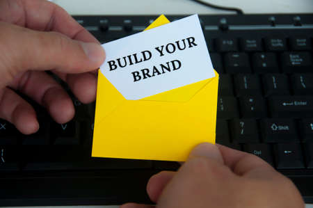 Hand holding with notepad in yellow envelope with text - Build your brand. With keyboard monitor...