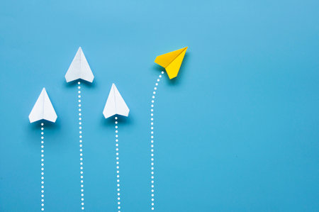Yellow paper plane leaving other white airplanes on blue background with customizable space for text or ideasの写真素材