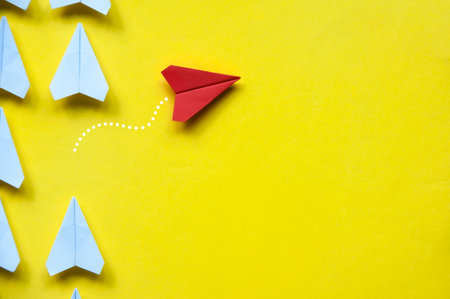 Top view of red paper airplane origami leaving other white airplanes on yellow background with customizable space for text or ideas. Leadership skills concept and copy spaceの写真素材