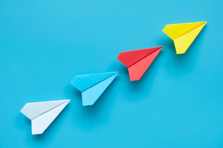 Yellow paper airplane origami leading other multicolored paper airplanes on blue background...