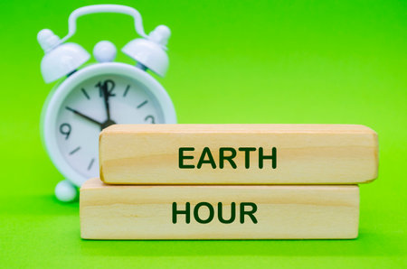 Wooden blocks with Earth Hour text placed on green surface with alarm clock background.の写真素材