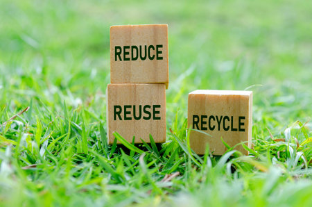 Wooden cubes labeled REDUCE, REUSE and RECYCLE on grass. Promoting sustainability.