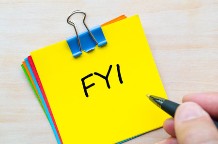 FYI text handwritten on yellow notepad with silver clip on wooden desk surfaceの写真素材