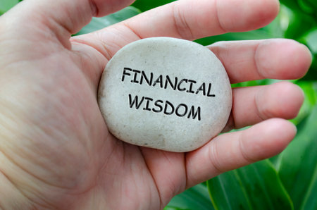 Hand holding a white stone with FINANCIAL WISDOM text engraved on it, symbolizing life purpose of stability and responsible livingの写真素材