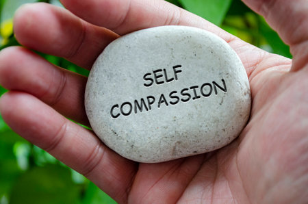 Hand holding a white stone with SELF COMPASSION text engraved on it, signifying life purpose of kindness toward oneself and healingの写真素材