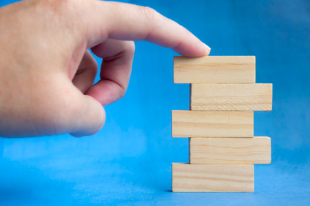 Hand holding wooden block above stacked pieces on blue background showing building and growth concept with customizable space for text or ideasの写真素材
