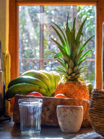 A glass and a clay cup in front of a wooden tray with bananas, a pineapple and some apples near a window in a farm house in central Colombia.の写真素材