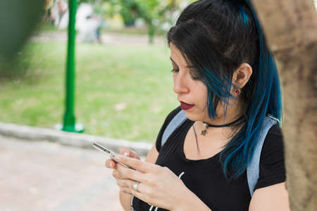 Beautiful young blue haired college girl looking at her social networks concentrated, in a park leaning on a treeの写真素材