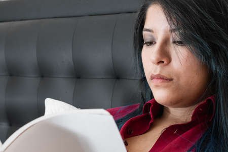 blue haired serious woman sitting on the bed reading a book concentrated