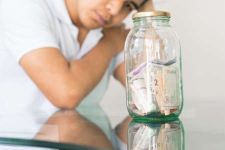 young guy crestfallen looking at his savings in a glass jar. young man next to his personal finances. bored student with a hand on his head. savings and finance conceptの写真素材
