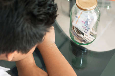 Disappointed young man looking at his savings, glass jar with money, Colombian banknotes, finance....
