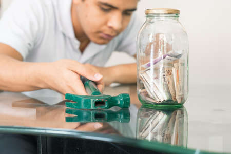 boy with hammer in hand next to glass jar full of banknotes. young man next to his personal finances. thrifty boy. savings and finance concept. man bad economicallyの写真素材