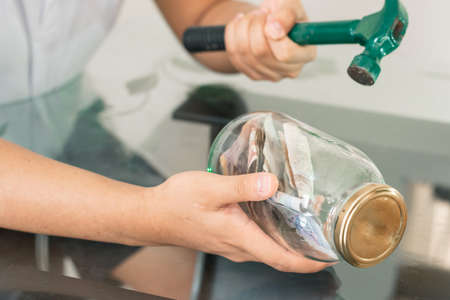 Young man with hammer in hand breaking his piggy bank, glass jar full of banknotes. savings and finance concept. Colombian economyの写真素材