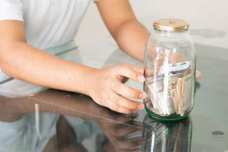 child's hand collecting their savings in a glass jar. young man next to his personal finances. thrifty boy. savings and finance conceptの写真素材