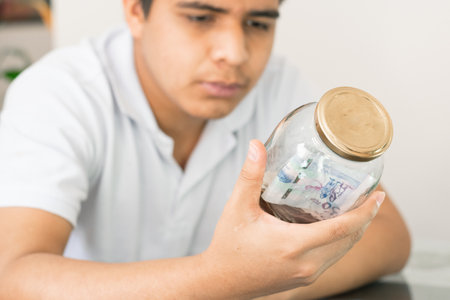 serious guy with glass jar full of banknotes in hand. young man collecting personal finances from him. thrifty boy. savings and finance concept. man looking at his savingsの写真素材