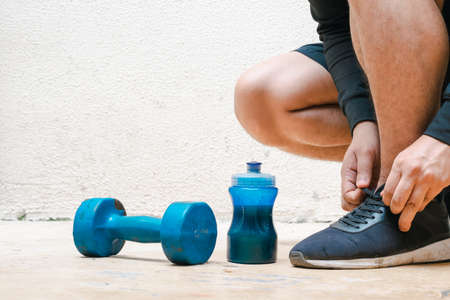man tying his shoes next to dumbbell and bottle with blue water, before exercising. exercise at home, white background. fitness concept. health concept, space for advertising textの写真素材