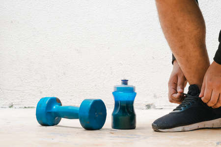 man tying his shoes next to dumbbell and bottle with blue water, before exercising. exercise at home, white background. fitness concept. health concept, space for advertising textの写真素材