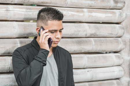 young latino university student talking on his cell phone with a serious face, leaning on a guadua hut, entrepreneur or influencer with his cell phone in his hand doing business. technological conceptの写真素材