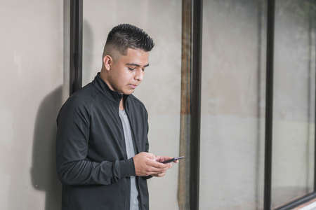 young entrepreneur looking at social networks on his cell phone, leaning against a glass window,...