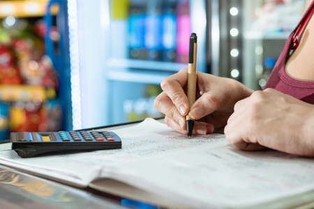 latina woman writing with her eco-friendly cardboard pen on a large notebook and doing calculations on her calculator about the sales and statistics in her grocery store. business conceptの写真素材