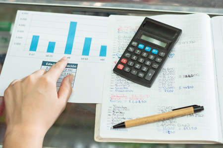 top view of a girl's hand pointing to her business statistics chart. next to her an eco-friendly cardboard pencil and a small black calculator, on top of a large notebook.の写真素材