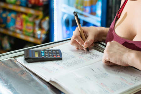 latina woman writing with her eco-friendly cardboard pen on a large notebook and doing calculations on her calculator about sales and statistics in her grocery store. on top of a display caseの写真素材