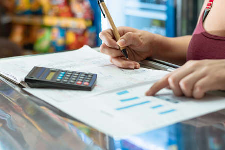 latina woman pointing to her business statistics chart. using her small calculator and cardboard eco-friendly pen on a large notebook. doing calculations on her grocery store sales.の写真素材
