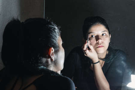 beautiful latina woman in front of the mirror making up her eyes. girl looking at herself in the mirror painting her eyebrows at home with a makeup brush. beauty, cosmetics and handicraft concept.の写真素材