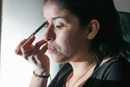 beautiful latina woman with green hair, making up her eyes with a black brush very concentrated in her makeup. making up her eyes with a red eye shadow beauty conceptの写真素材