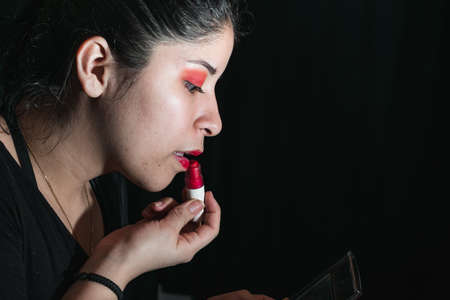 latina woman using a lipstick to paint her lips red, opening her mouth a little to apply the lipstick. her eyes are painted with a red shadow. black background. looking at herself in the mirror.の写真素材