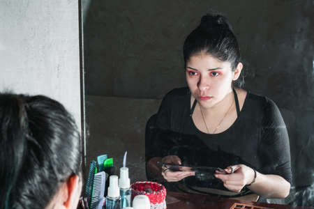beautiful latin woman in front of the mirror, with her gaze fixed on her eyes that she has just made up, observing with a serious look if her make up is ok. concept of beauty and cosmeticsの写真素材