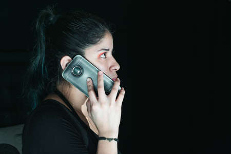 beautiful latina woman with green hair receiving an extortion call, with her cell phone in her hand. with her gaze upwards and serious look. with a black background. concept of technology and robbery.の写真素材