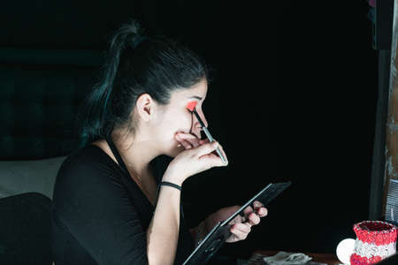 beautiful latina woman with green hair, making up her eyes with a red eye shadow. girl sitting in her room holding her palette of powdered colors. black background. natural beauty concept.の写真素材