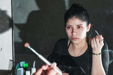 beautiful latina woman in front of the mirror, with her gaze fixed on her made-up eyes, observing if her make-up is well done. with a brush in her right hand. concept of beauty and cosmetics.の写真素材