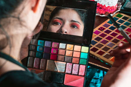 Beautiful Latina woman looking in the mirror of her eye shadow palette, with a palette of colored shadows in the background on top of her dressing table, girl doing her eye makeup with red color.の写真素材