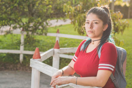 Beautiful young Latina college girl, waiting for her package next to a wooden fence, dressed in red and dyed blue hair, with a beautiful sunset in the background and a car driving away.の写真素材