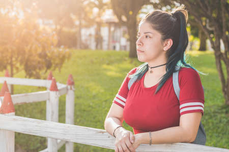 Beautiful young Latina college girl, pensive, dressed in red with blue dyed hair, standing next to a white fence, with a beautiful sunset in the background. concept of freedom.の写真素材