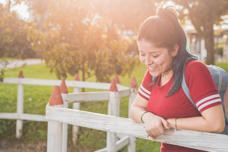 Beautiful college girl, standing next to a very happy wooden fence, smiling and blushing, looking down at the ground, dressed in red and with blue hair, with a beautiful sunset in the background.の写真素材