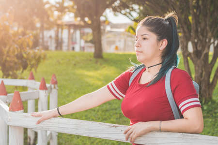 Beautiful young woman student, standing next to a wooden fence, very serious and worried about her life project, dressed in red and with blue dyed hair, with a beautiful sunset in the background.の写真素材